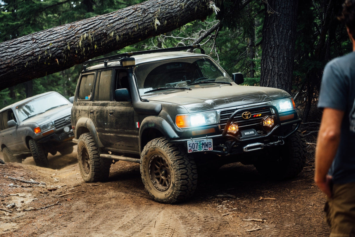 Dark 80 Series Land Cruiser with its lights on driving under a fallen tree on a dirt trail, another 80 behind it