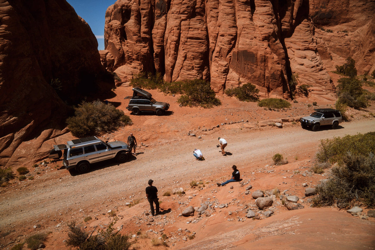 Three overland trucks and their crew spread along a red dirt road below sandstone cliffs