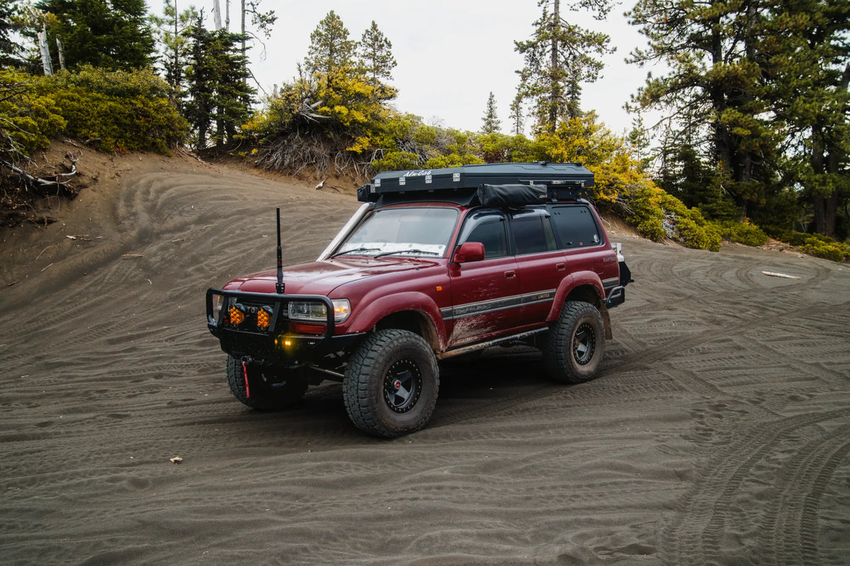 Red 1990 Land Cruiser HDJ81 with an Alu-Cab pop-top, parked on black sand below a bank of pines