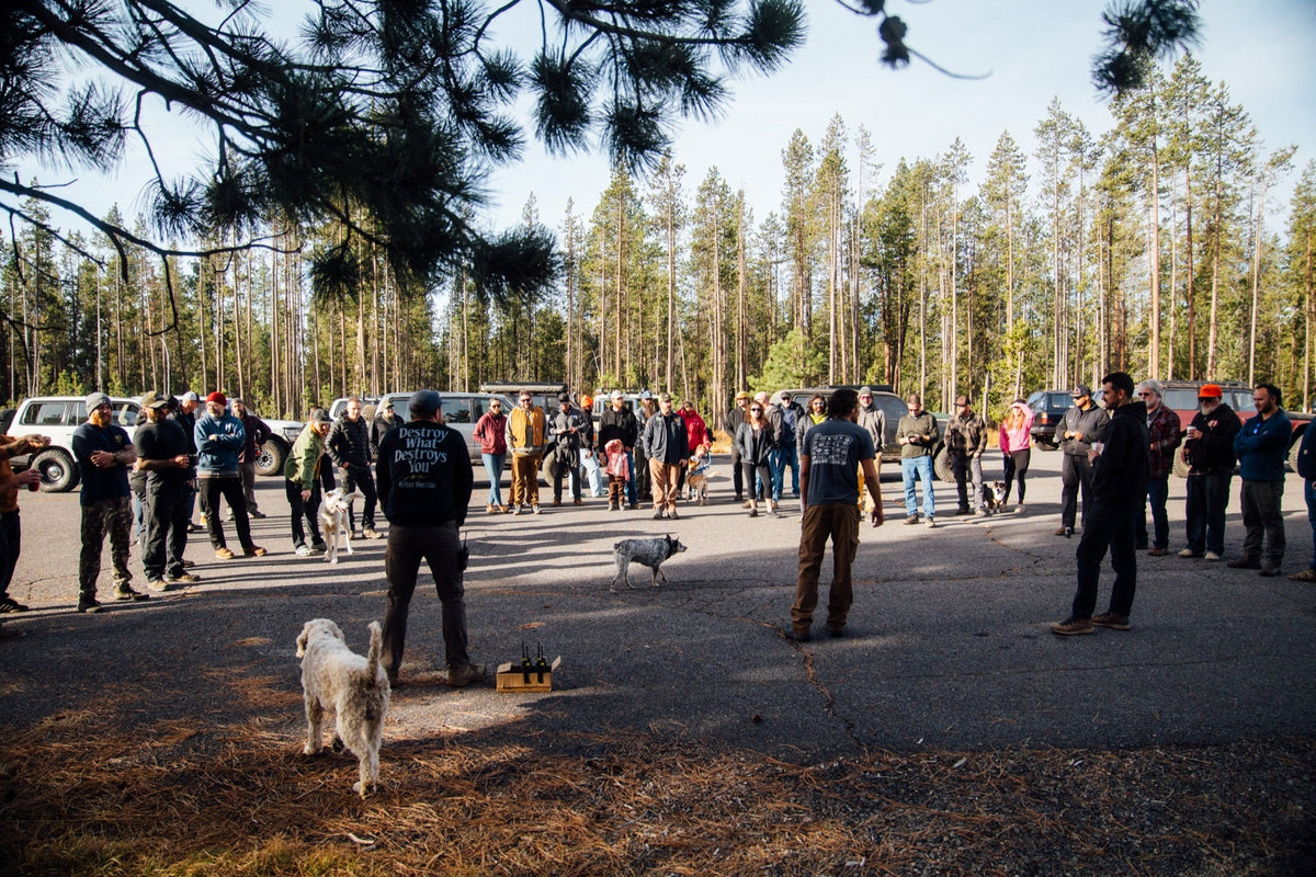 About thirty people at the Eighties on Edison drivers meeting in a gravel lot among pines, a dog wandering through