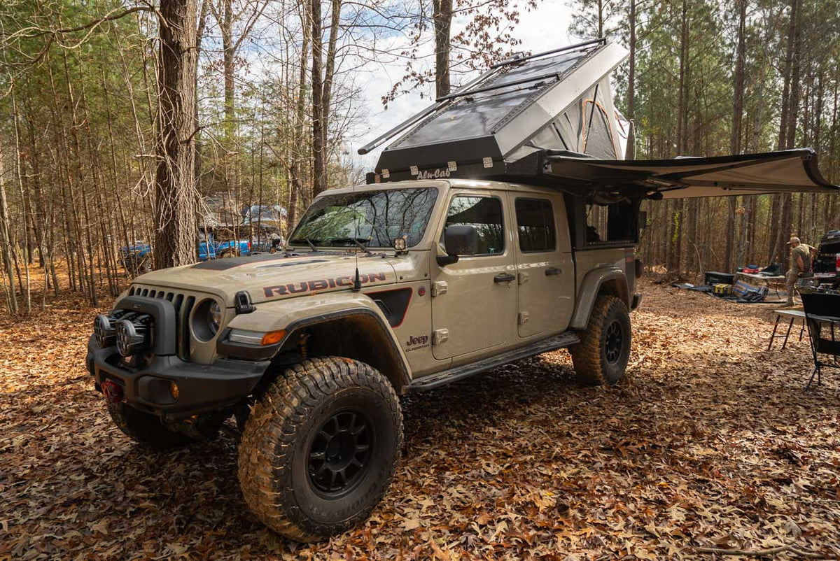 A Jeep Gladiator with a bed camper parked in a wooded camp