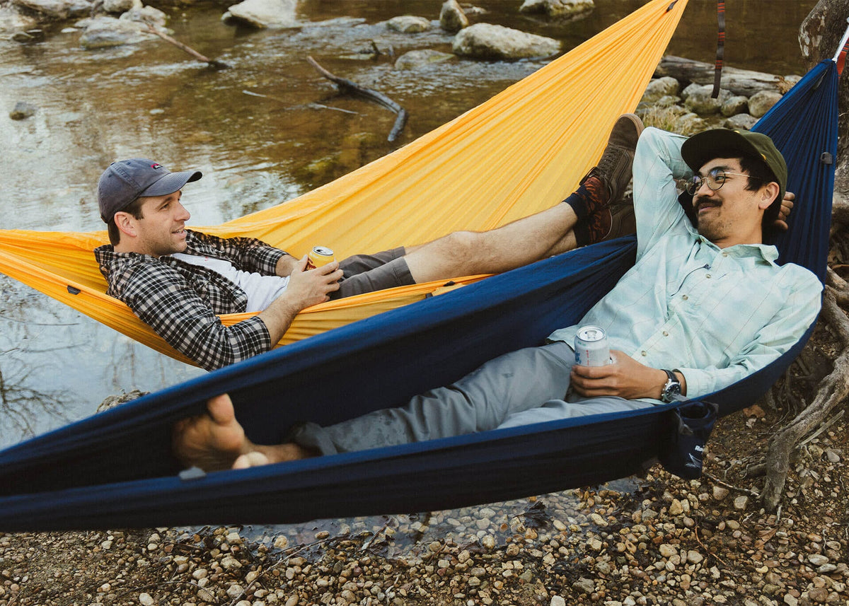Two people in Roo Single hammocks in yellow and navy, hung low over a creek