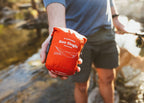 The Roo Single packed in its orange stuff sack, held in one hand beside a creek