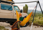 Two people in a yellow hammock hung from the Outpost on the back of a tan van, cactus behind