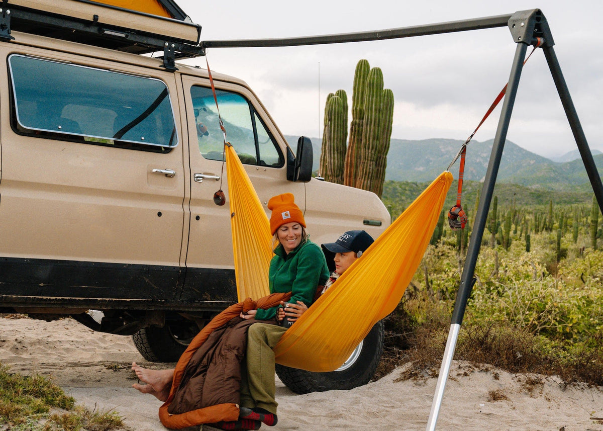 Two people in a yellow hammock hung from the Outpost on the back of a tan van, cactus behind
