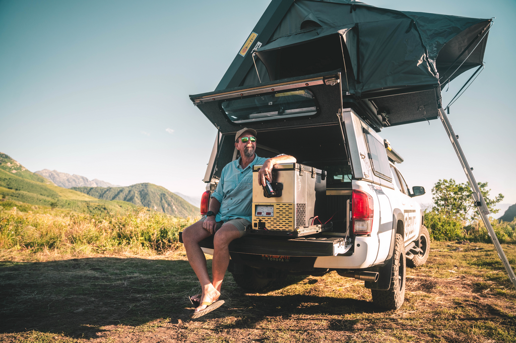 National Luna 35L dual zone fridge freezer on a slide at a pickup's tailgate, a person seated beside it, rooftop tent above