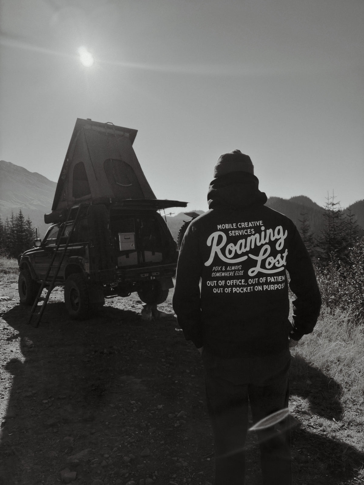 The Mobile Creative Services hoodie from behind, back print showing, worn beside an 80 Series Land Cruiser with its rooftop tent open on a mountain road