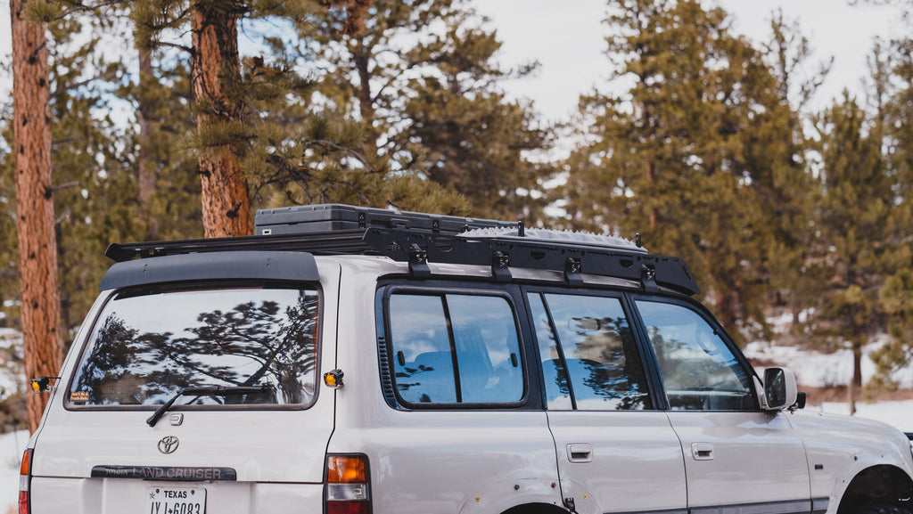 White 80 Series Land Cruiser from the rear quarter with a La Sal rack and a roof box, pines and snow